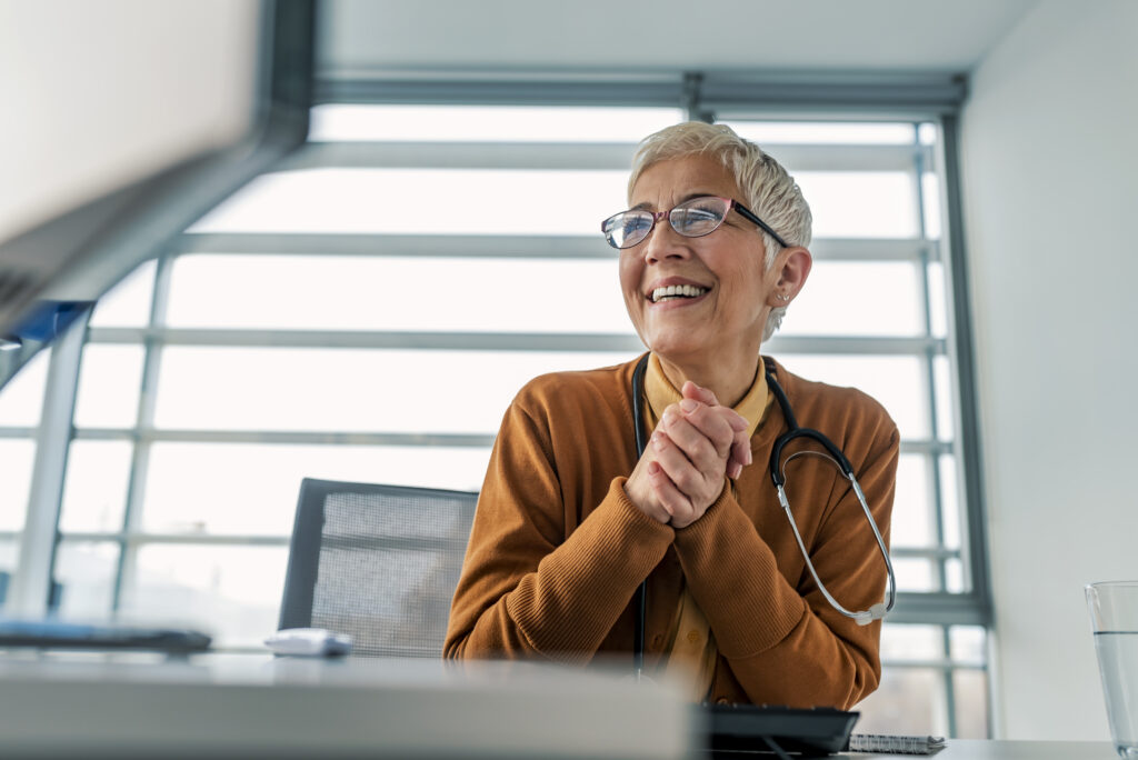 Photo of happy Senior female doctor, working at desk, using PC computer. Looking away with smile on her face. Mature positive gar hair female doctor working on medical expertise while sitting at desk in front of computer. Elderly businesswoman working at her desk at the office reading through notes with stethoscope around her neck.